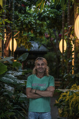 Portrait of mature man in t-shirt, jeans standing on the green street. Long grey hair. Chinese lanterns. Evening. Trees on the background.