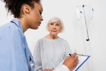 Elderly patient in gown looking at blurred african american nurse writing on clipboard