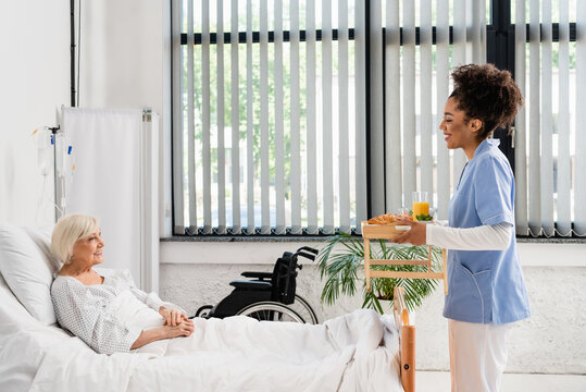 Side View Of Smiling African American Nurse Holding Food On Tray Near Senior Patient