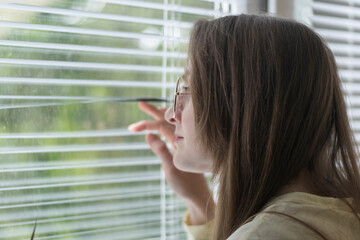 Young woman looks out window through the blinds. Girl with glasses looks at street through blinds.