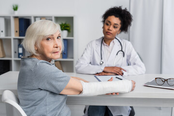 Senior woman with plaster bandage on arm looking at camera near blurred african american doctor