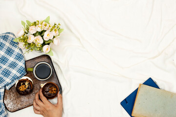 hand  holding one chocolate macaron ,agenda book or notebook and cup of hot coffee with macarons, flowers pot and pillow on white bed