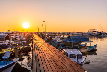 Obraz premium tranquil sunrise in fishing docks with sunny pier , leading far to sea and golden sun with sailing boats moored around