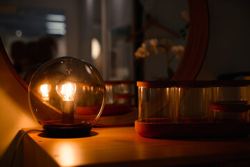 Home evening interior close up. Luminous light bulb inside glass ball, standing on wooden chest of drawers, indoors. Selective focus of decorative lamp in trendy lampshade.