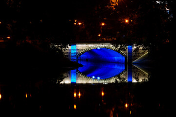 Night photo of a pedestrian bridge over a canal. The bridge is illuminated with blue lights that...