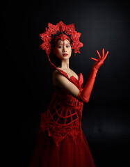 Full length  portrait of beautiful young asian woman wearing red corset, long opera gloves and ornate gothic queen crown. Graceful posing isolated on a dark studio background.