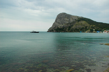 Fototapeta premium The boat sails along the rocky coast of the Black Sea among rocks and mountains. Natural landscape and seascape. People travel and relax on the ship. Clear green water, algae. Stone rocks. Crimea.