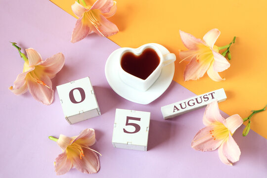 Calendar for August 5 :the name of the month of August in English, cubes with the number 05, a cup of tea in the shape of a heart, daylily flowers on a yellow-lilac background, top view