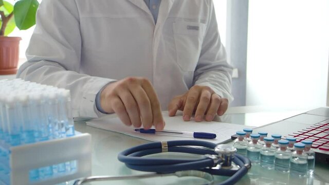 Doctor works in front of a laptop monitor. He swipes the papers, puts down his pen and takes off his glasses. Looks like he's got research problems.