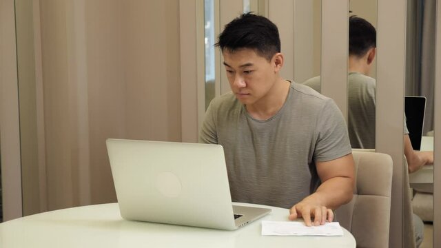 Serious asian male accountant sitting in home office do work distantly managing company budget, count financial plan use calculator calculates expenses typing writing digits results into application.