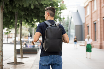 Rear view man walking in city with black leather backpack on his shoulders - obrazy, fototapety, plakaty