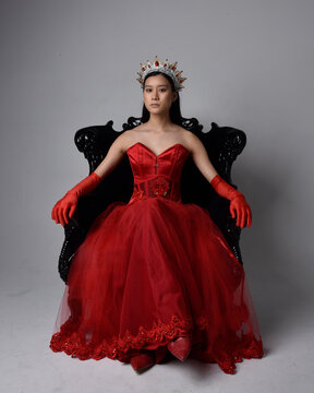 Full length  portrait of beautiful young asian woman wearing red corset, long opera gloves and ornate gothic queen crown. Graceful posing while seated on chair, isolated on studio background.