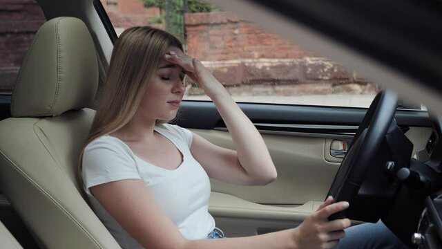 The stressed blonde woman driver is sitting inside her car. Sad young girl is having headache has to make a stop after driving car in traffic jam on rush hour. Exhausted, overworked driver concept.