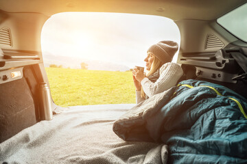 Girl resting in her car. Woman hiker, hiking backpacker traveler camper in sleeping bag, drinking hot tea and relaxing on top of mountain. Health care, authenticity, sense of balance and calmness.