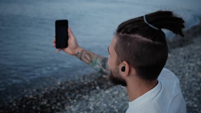 Remote work and communication on vacation. Successful businessman. Caucasian man with dreadlocks in white shirt is sitting on beach with ear headphones and chatting via facetime on his phone.