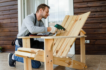 Young carpenter assembling wooden adirondack chair with electric screwdriver or drill. Small...