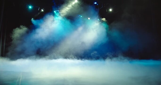 Rotating beams of stage light shine through the heavy white smoke, they form the backdrop for the concert. The background is painted blue and white.