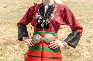 Old traditional women's clothing. A young girl - a woman in Bulgarian folk costume. Silver ornaments, red robe and silver belt. Women's clothing up close.
