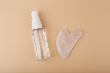 Flat lay with massage gel or oil in transparent bottle and heart shaped guasha pink quartz crystal on beige background. Concept of facial massage, beauty and self treatment