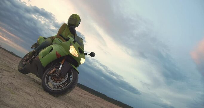 A sexy and attractive young woman in a helmet sits on a high-speed green motorcycle. She's wearing a matching T-shirt. The girl is all smeared with dry paint. A beautiful view of the sky is visible