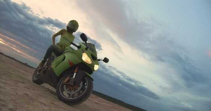 An attractive young woman sits on a sporty green motorcycle. She is wearing the same color T-shirt and safety helmet. She leans back and almost lies down. In the background, a cloudy sky and sand