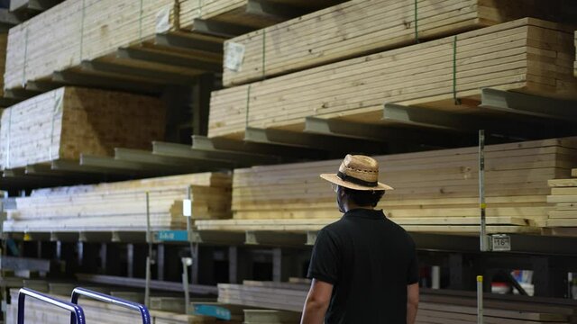 Man walks down aisle with stacks and piles of lumber, wood, and sheets of plywood in a big box hardware store.