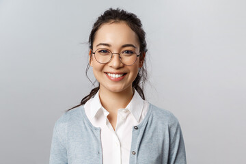 Close-up portrait of attractive, friendly-looking asian female office worker, employee or teacher in glasses, smiling broadly camera with enthusiastic attitude, stand grey background - Powered by Adobe