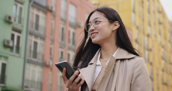Happy Young Asian Girl Student in Eyeglasses walking while Browsing Smartphone. Smiling Chinese Woman using Mobile Applications and chatting on Social Media. Wears Trendy Coat. Networks. Notifications