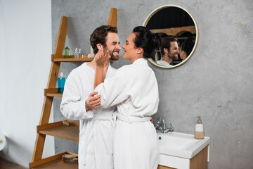 joyful woman applying face cream on nose of smiling boyfriend in bathrobe