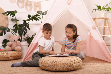 Cute little children reading book near toy wigwam at home © New Africa