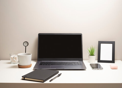 Picture of laptop on a white table with a mug a clock a small bush a notebook a phone a picture frame and a headphone case