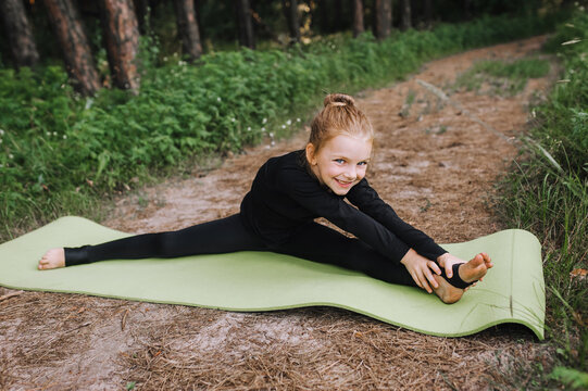 A beautiful girl, a child, a professional gymnast in a black suit trains diligently, stretches in a twine pose in the woods in nature on a green long rug. Sports photography.