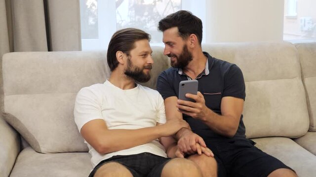 Guys Smile and Laugh in Love at Home. Cute Male Homosexual Gay Couple Spend Time Together on the Couch. Men Surfing Social Media on Smartphone. LGBTQI, Pride Event, LGBT Pride Month, Gay Pride Symbol.