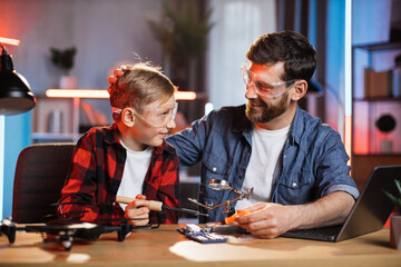 Positive caucasian father teaching his little son how to fix display card from modern laptop. Man and boy working with soldering iron at home.
