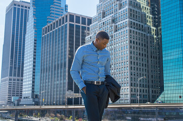 A young black businessman is standing in a business district, looking down and into deeply...