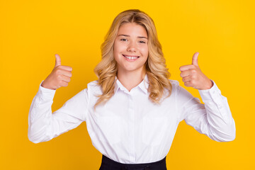 Photo of cheerful schoolgirl raise two thumbs up beaming shiny smile wear white shirt isolated yellow color background