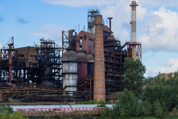 Fototapeta na wymiar The building of the old iron-smelting plant against the background of the cloudy sky. Smoked blast furnaces and metal structures, industrial landscape.