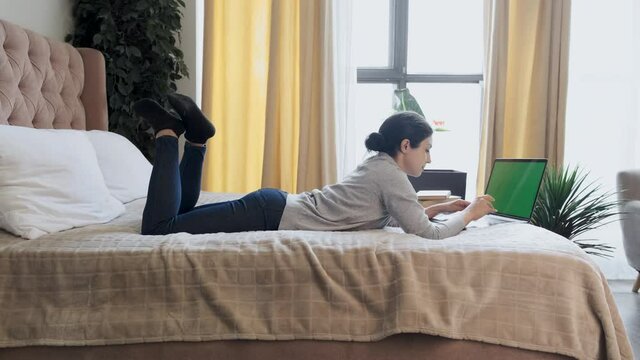 A Young Indian Thoughtful Woman Is Typing on a Keyboard, Working Online at a Computer with a Green Screen, Lying on a Bed in a Bright Room