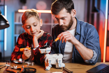Handsome man and cute boy in casual clothes using screwdrivers for adjustment of interactive toy. Father with son sitting together at table and fixing robot dog.