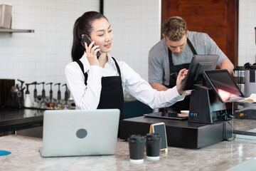 woman barista talking with smartphone and take orders online to customer with laptop computer