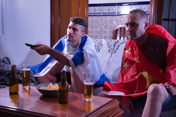 Two men one mature and one young watching sports competitions on television, changing channels with the command with flags of China and Israel.