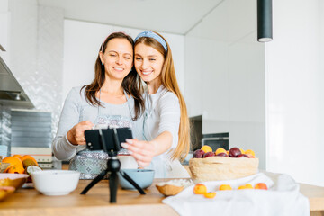 Happy fresh middle age woman with her adult daughter having a video call over smart phone at home. Smiling housewives cooking at kitchen during online video call with her family.