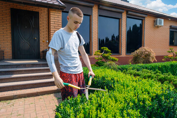 Young caucasian man with an amputated arm and a prosthesis is trimming bushes in a garden in his...