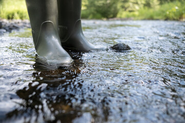 Male feet in waterproof rubber boots walk along the river, against the backdrop of greenery, on a...
