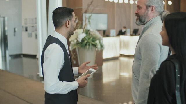 Slowmo shot of polite male concierge welcoming modern guest couple in hotel lobby, escorting them to reception desk