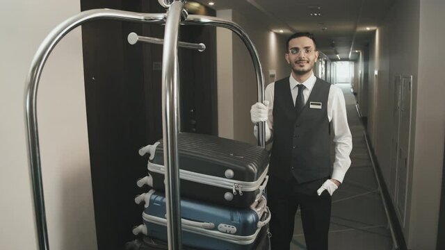 Medium arc shot portrait with slowmo of professional male porter in gloves and uniform posing for camera with baggage cart in hotel corridor