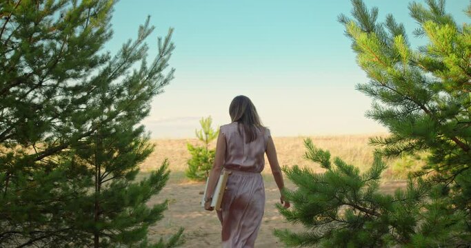 View from the back of a young woman in a pink dress running along the sand between coniferous trees.