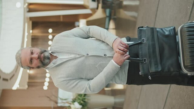 Vertical medium slowmo portrait of confident mature businessman in smart casualwear standing with luggage in posh modern hotel lobby looking at camera