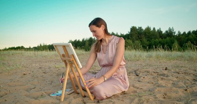 Young woman sits in nature near an easel with a palette and paints a landscape.