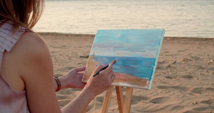 A woman sits on the sand by the sea and paint a seascape on the canvas with a brush.